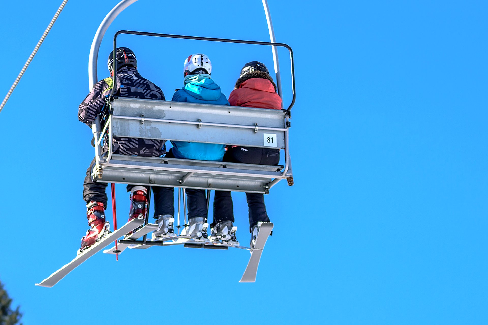 Boot Room Ski Shop. Looking up at three colorful chairlift riders — boots and skis against blue sky.