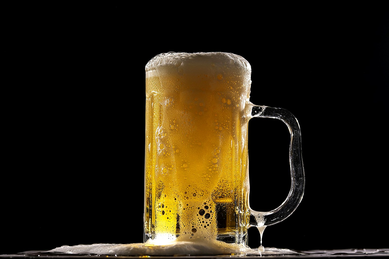 Backlit golden beer stein overflowing with foam on a dark surface.