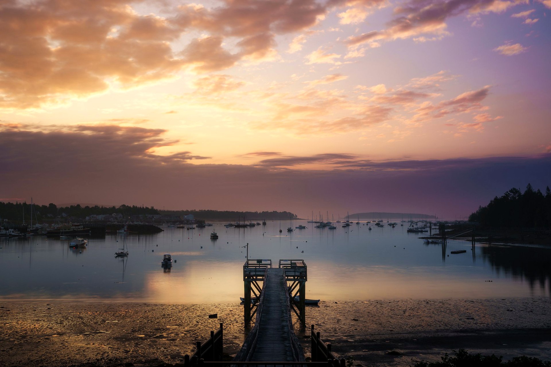 Long wooden pier into a calm harbor at golden sunset with boats at anchor.