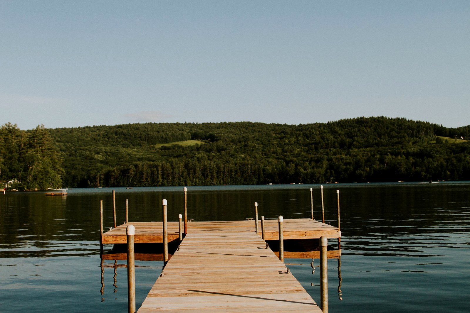 Long wooden dock stretching into a calm lake toward forested hills under pale sky.